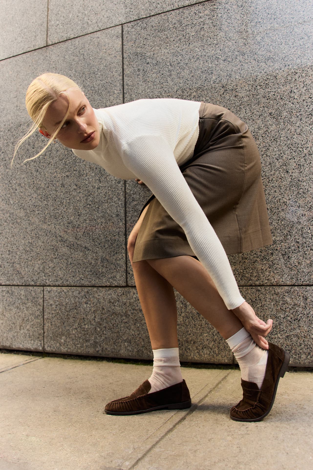 Betts campaign image of a model in a cream knit and brown suede loafers photographed against a city building